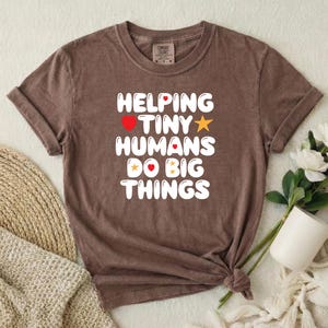 May include: A brown short-sleeved t-shirt featuring the white text HELPING TINY HUMANS DO BIG THINGS, accented with small red hearts and yellow stars. The shirt has a crew neckline and a tag at the inner collar indicating the brand Comfort Colors.