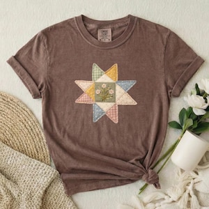 Könnte beinhalten: Ein braunes Kurzarm-T-Shirt mit einer zentralen sternförmigen Grafik, die aus verschiedenen gemusterten Stoffstücken in gedeckten Gelb-, Blau-, Rosa- und Grüntönen besteht, mit einer kleinen Blumenillustration in der Mitte des Sterns.