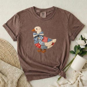 Puede incluir: Una camiseta marrón de manga corta con un gráfico de un ganso hecho de varios parches de tela. El ganso lleva un lazo de cuadros azules y blancos y tiene un ala de cuadros rojos y blancos. La camiseta tiene cuello redondo y una etiqueta.