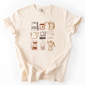 May include: A cream-colored t-shirt featuring a brown graphic print of various coffee-related items, including a bag labeled COFFEE, a cup, a French press, a kettle, a pour-over dripper, an espresso machine, a drip coffee maker, and a moka pot.