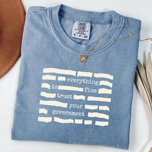 Può includere: Una t-shirt blu a maniche corte con scollo rotondo e una stampa grafica bianca. Il testo "everything is fine trust your government" è disposto in righe separate da barre bianche orizzontali. Una collana color oro è appoggiata sulla maglietta.