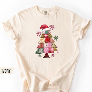 May include: An ivory short-sleeve t-shirt featuring a central graphic of a Christmas tree constructed from various patchwork fabric patterns in red, pink, green, and gold, topped with a red and white Santa hat and surrounded by small snowflakes.