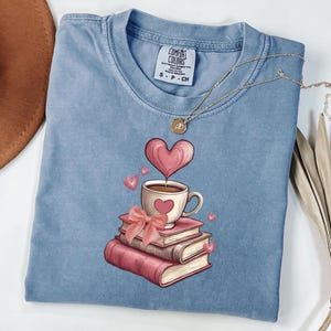May include: A blue short-sleeved t-shirt with a Comfort Colors label at the neck. The graphic features a stack of three books, a coffee cup with a heart, a pink bow, and floating hearts. A gold-toned necklace rests on the fabric.