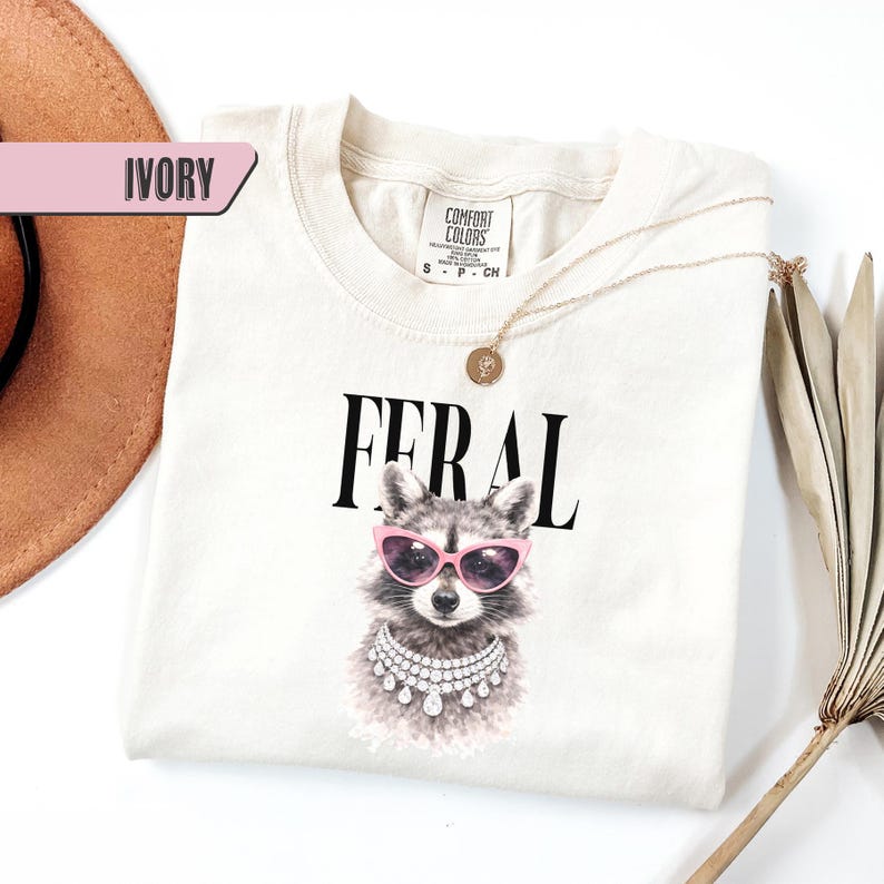 May include: Ivory-colored t-shirt with the word "FERAL" in black block letters. The shirt features a raccoon wearing pink sunglasses and a diamond necklace. A gold necklace with a pendant is draped across the shirt.