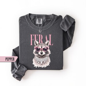 May include: Dark grey long-sleeve shirt with a graphic of a raccoon wearing pink sunglasses and a diamond necklace. The word "FERAL" is printed above the raccoon in pink letters. The shirt is labeled "PEPPER".
