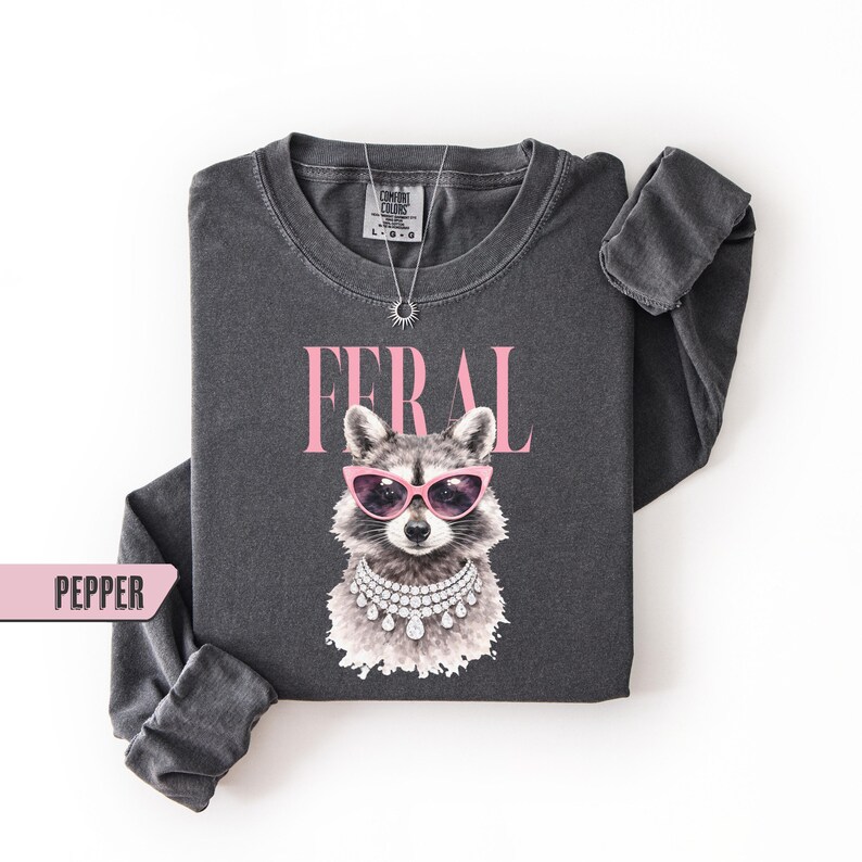 May include: Dark grey long-sleeve shirt with a graphic of a raccoon wearing pink sunglasses and a diamond necklace. The word "FERAL" is printed above the raccoon in pink letters. The shirt is labeled "PEPPER".