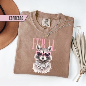 May include: Espresso-colored t-shirt with a raccoon graphic wearing pink sunglasses and a jeweled collar. The word "FERAL" is printed above the raccoon in pink letters. A silver necklace and a brown hat are also visible.