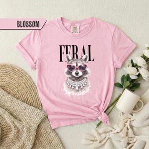 May include: Pink t-shirt with the word "FERAL" in black block letters. The shirt features a graphic of a raccoon wearing pink sunglasses and a jeweled necklace. The word "BLOSSOM" is in a pink banner in the upper left corner.