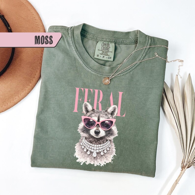 May include: A moss green t-shirt featuring a raccoon wearing pink sunglasses and a jeweled necklace. The word "FERAL" is printed above the raccoon in pink letters. A gold necklace is draped on the shirt.