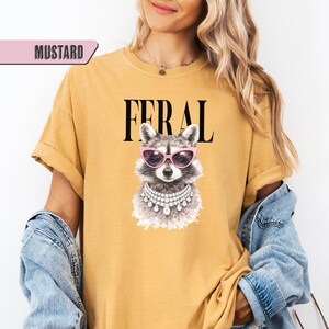 May include: Mustard-colored t-shirt with the word "FERAL" in black block letters. The shirt features a graphic of a raccoon wearing pink sunglasses and a pearl necklace. The shirt is a casual style.