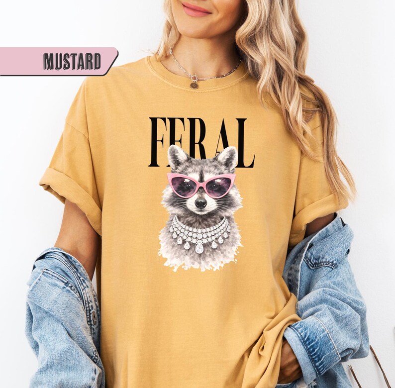 May include: Mustard-colored t-shirt with the word "FERAL" in black block letters. The shirt features a graphic of a raccoon wearing pink sunglasses and a pearl necklace. The shirt is a casual style.