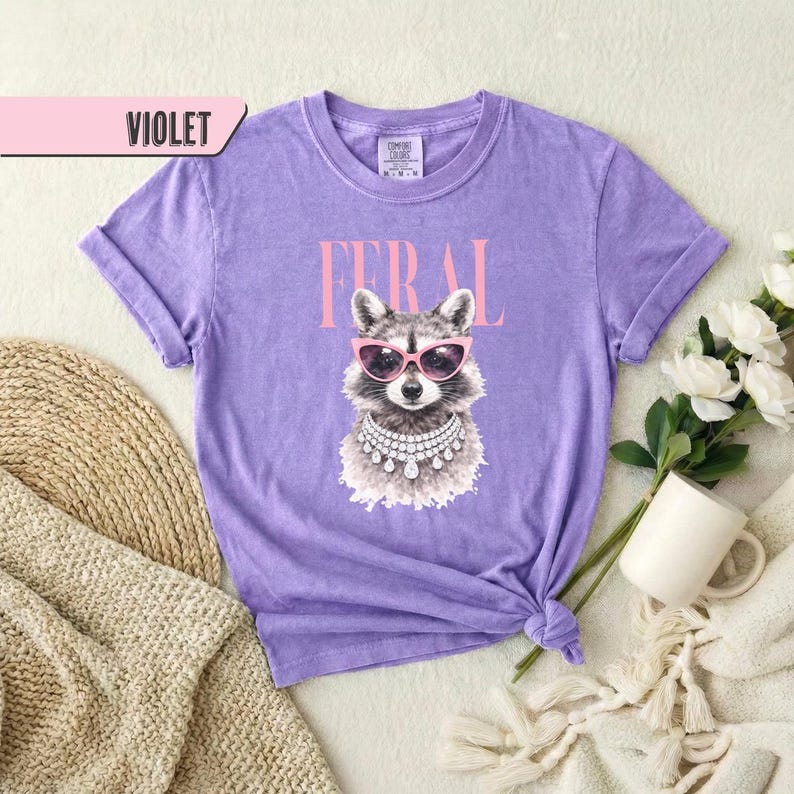 May include: Violet-colored t-shirt with a graphic of a raccoon wearing pink sunglasses and a pearl necklace. The word "FERAL" is printed above the raccoon in pink letters. The word "VIOLET" is in a pink rectangle in the upper left corner.