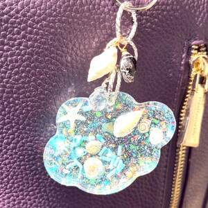 Könnte beinhalten: Ein wolkenförmiger Schlüsselanhänger mit einem silberfarbenen Ring. Der Schlüsselanhänger ist mit Glitzer, Muscheln und kleinen Seesternen gefüllt. Er hat auch einen kleinen Totenkopf-Anhänger und ein paar klare Kristallperlen. Der Hintergrund ist eine lila Ledertasche.