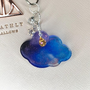 Könnte beinhalten: Ein wolkenförmiger Schlüsselanhänger mit blauem und lila Galaxiemuster. Der Schlüsselanhänger hat einen silbernen Verschluss und einen kleinen Stern- und Kristallanhänger. Ein goldfarbener Tropfenanhänger hängt an der Wolke.