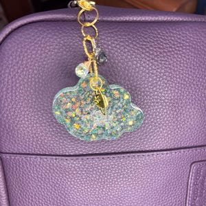Könnte beinhalten: Ein wolkenförmiger Schlüsselanhänger mit einem goldfarbenen Verschluss und einer Kette. Der Anhänger ist mit Glitzer und kleinen bunten Flecken gefüllt. Ein kleiner goldener Blattanhänger hängt an der Wolke. Der Anhänger ist an einer lila Tasche befestigt.