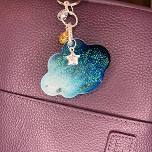 Könnte beinhalten: Ein wolkenförmiger Schlüsselanhänger mit einem silbernen Sternanhänger und einer goldfarbenen Tränenperle. Die Wolke ist eine Mischung aus blauem und grünem Glitzerharz. Der Schlüsselanhänger ist an einem silbernen Schlüsselring befestigt.