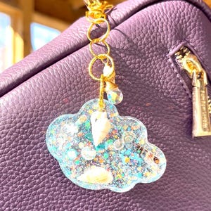Könnte beinhalten: Ein wolkenförmiger Schlüsselanhänger mit einem goldfarbenen Verschluss und einer Kette. Die Wolke ist mit blauem Glitzer und kleinen weißen Objekten gefüllt. Der Schlüsselanhänger ist an einer lila Ledertasche befestigt.