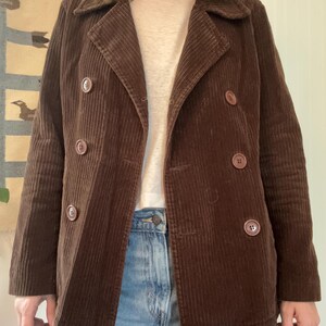 May include: A brown corduroy jacket with a double-breasted design. The jacket features large, round buttons and a notched collar. The corduroy fabric has vertical ridges, and the jacket is worn over a light-colored shirt and blue jeans.