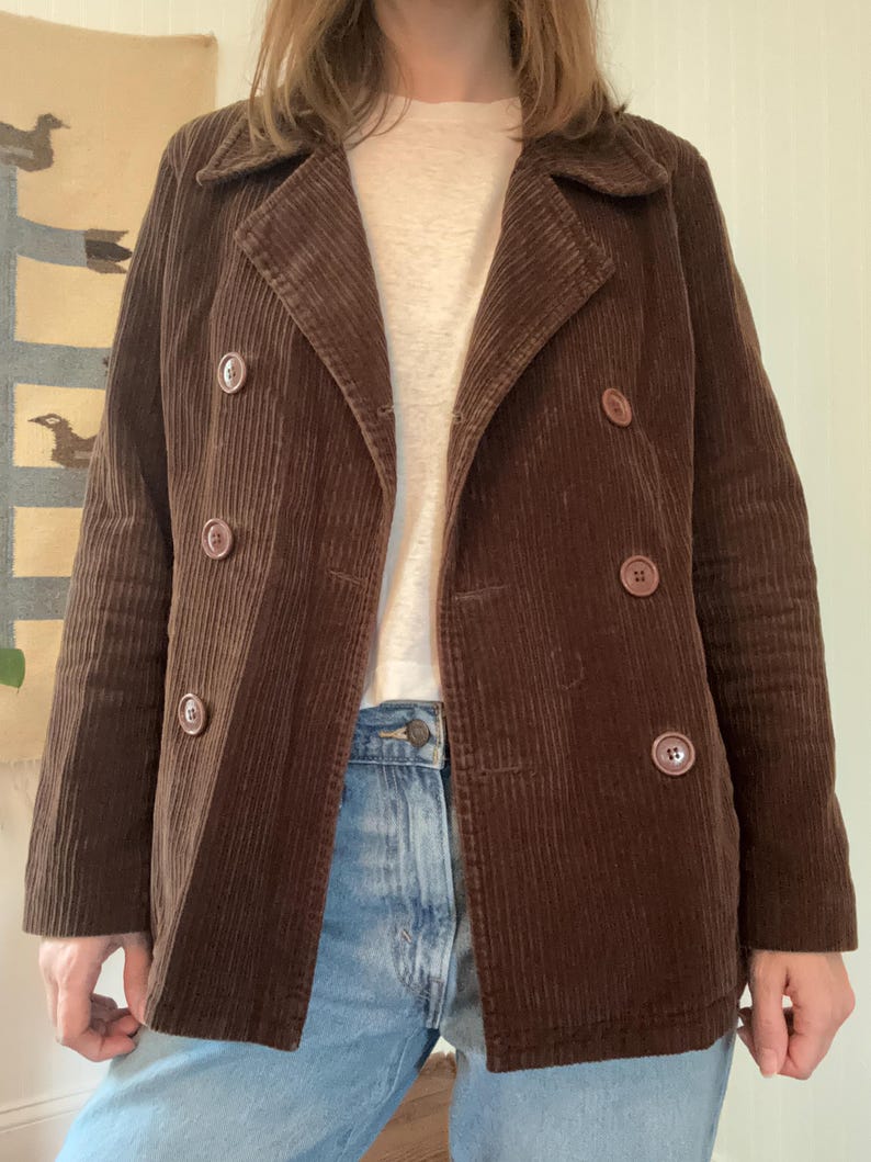 May include: A brown corduroy jacket with a double-breasted design. The jacket features large, round buttons and a notched collar. The corduroy fabric has vertical ridges, and the jacket is worn over a light-colored shirt and blue jeans.