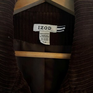 May include: A close-up of a brown corduroy jacket hanging on a wooden hanger. The jacket has a label that reads "IZOD" and "100% Cotton Made in El Salvador". The jacket is a small size.