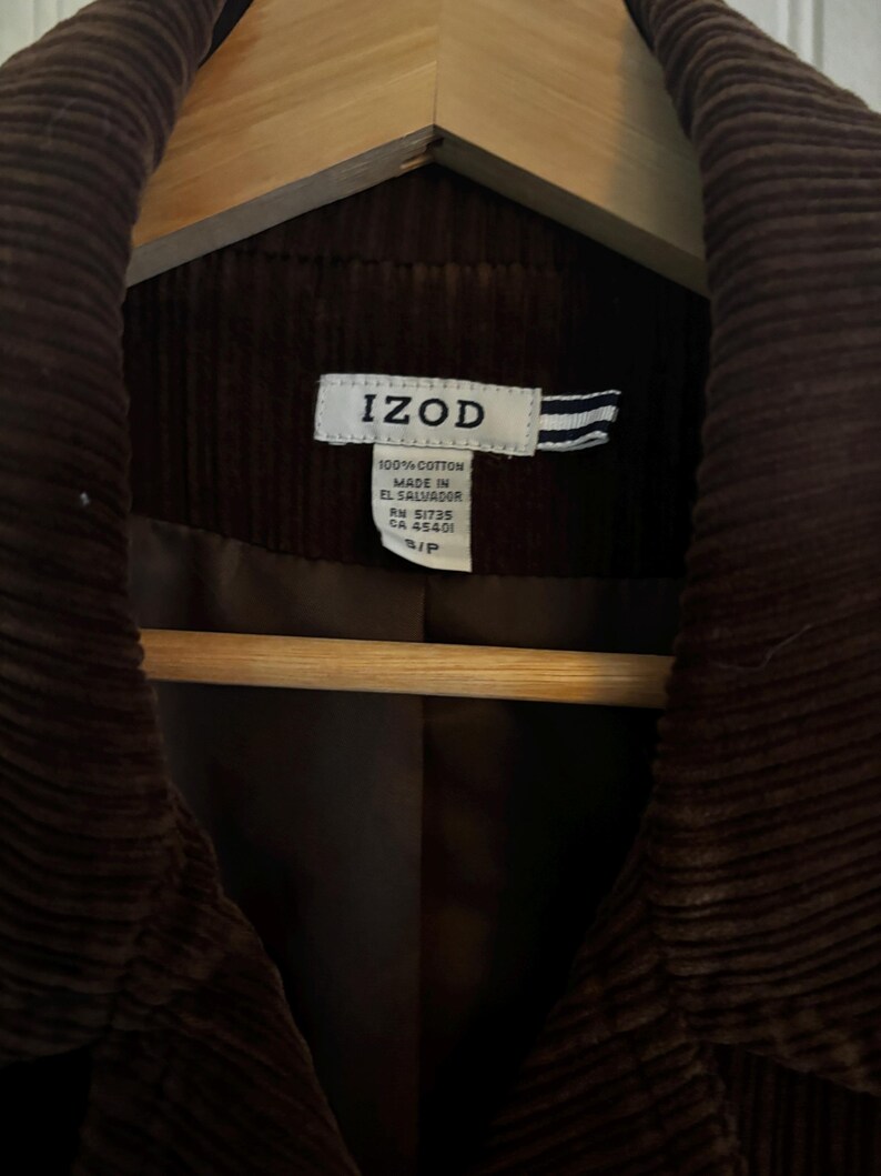 May include: A close-up of a brown corduroy jacket hanging on a wooden hanger. The jacket has a label that reads "IZOD" and "100% Cotton Made in El Salvador". The jacket is a small size.