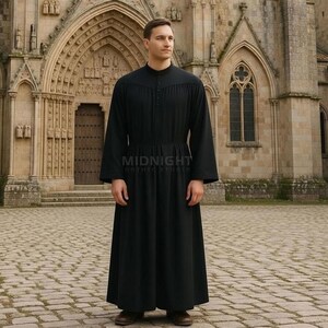 Op de afbeelding: Een zwarte, lange mouwen jurk met een hoge kraag en knoopdetails. De jurk heeft een getailleerde taille en een zwierige rok. De tekst "MIDNIGHT GOTHIC STUDIO" is op de voorkant gedrukt. De persoon staat voor een stenen gebouw.
