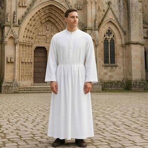 Op de afbeelding: Een witte, lange mouwen jurk met een hoge kraag en knoopdetails. De jurk heeft een geplooide taille en een zwierige rok, gedragen met donkerbruine schoenen. De achtergrond toont een stenen gebouw met gewelfde deuren en ramen.
