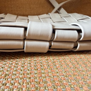 Pu&ograve; includere: Una borsa beige intrecciata con tracolla. La borsa presenta un motivo ripetuto di strisce intrecciate, creando una superficie strutturata. La borsa &egrave; appoggiata su un tessuto fantasia.