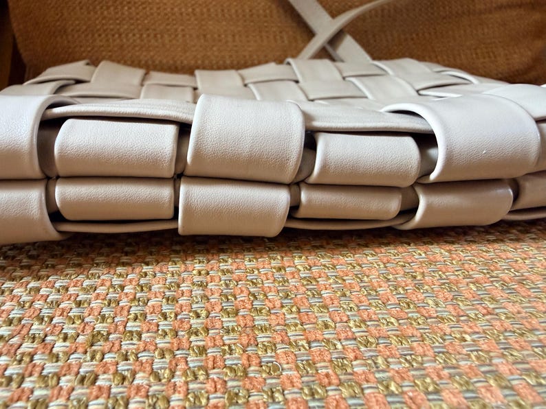 Pu&ograve; includere: Una borsa beige intrecciata con tracolla. La borsa presenta un motivo ripetuto di strisce intrecciate, creando una superficie strutturata. La borsa &egrave; appoggiata su un tessuto fantasia.