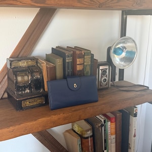 May include: A navy blue leather wallet with a gold-colored clasp sits on a wooden shelf, alongside vintage books, a retro camera, and an antique cash register. A large, silver-colored light fixture is also on the shelf.