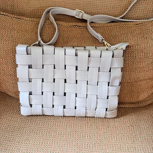 Pu&ograve; includere: Una borsa a tracolla grigio chiaro con un design intrecciato. La borsa ha una tracolla lunga e regolabile e una cerniera dorata. La borsa rettangolare &egrave; su una superficie marrone.
