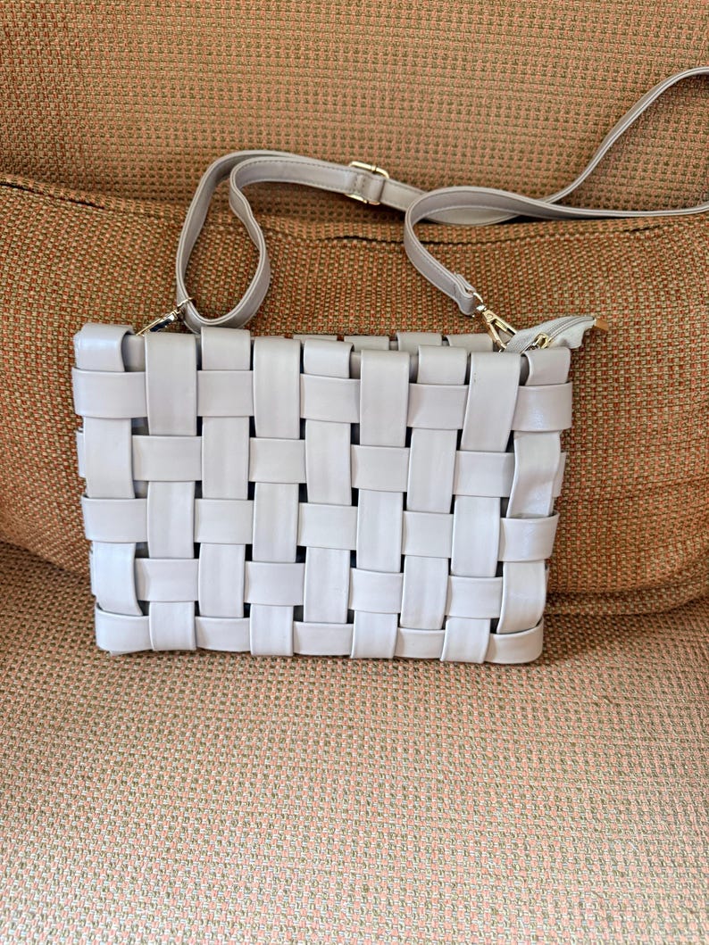 Pu&ograve; includere: Una borsa a tracolla grigio chiaro con un design intrecciato. La borsa ha una tracolla lunga e regolabile e una cerniera dorata. La borsa rettangolare &egrave; su una superficie marrone.