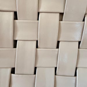 Pu&ograve; includere: Primo piano di un motivo intrecciato di colore crema chiaro. Il design presenta strisce rettangolari intrecciate per creare una superficie strutturata. L'intreccio &egrave; stretto e uniforme, con una sottile lucentezza sul materiale.