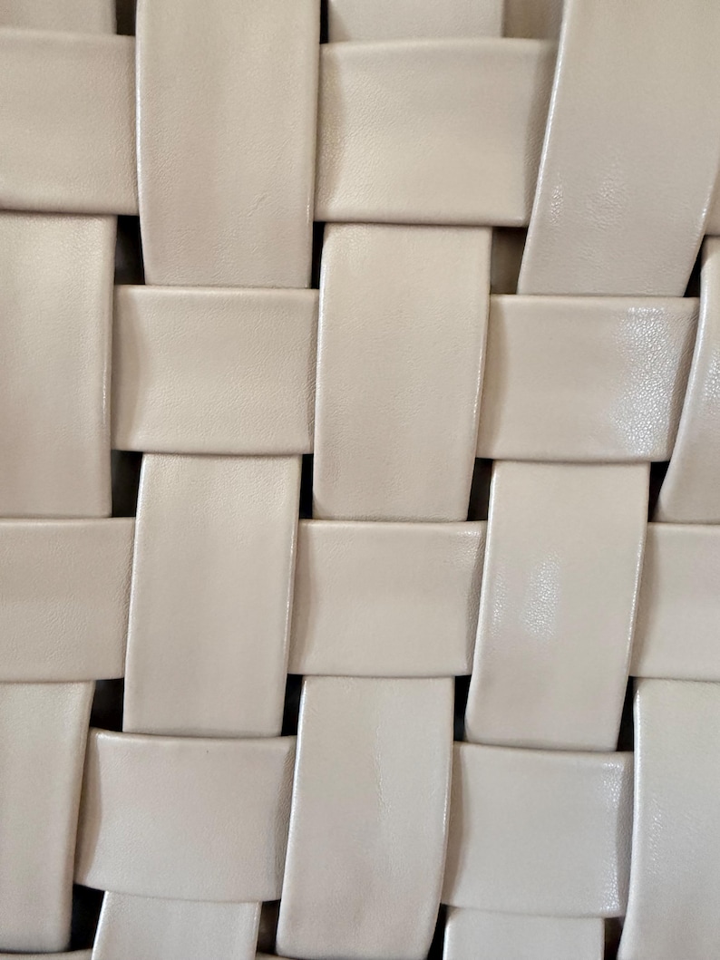 Pu&ograve; includere: Primo piano di un motivo intrecciato di colore crema chiaro. Il design presenta strisce rettangolari intrecciate per creare una superficie strutturata. L'intreccio &egrave; stretto e uniforme, con una sottile lucentezza sul materiale.
