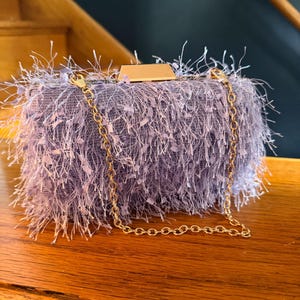 May include: A lavender clutch with a gold chain strap. The rectangular bag is covered in a fuzzy, textured material. A gold-colored clasp is visible on the top. The bag is sitting on a wooden surface.