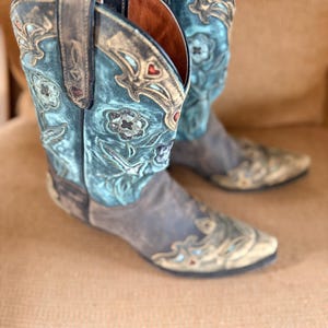 Op de afbeelding: Een paar turquoise en grijze cowboylaarzen met ingewikkelde bloemenborduurwerk. De laarzen hebben een puntige neus en een afgewerkte leren afwerking, met rode hartaccenten en een bruine zool.