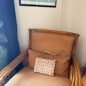 Pu&ograve; includere: Una borsa a mano beige chiaro intrecciata con una tracolla &egrave; appoggiata su un cuscino marrone. La borsa ha una forma rettangolare e una chiusura a cerniera. La borsa &egrave; su una sedia marrone con schienale e braccioli intrecciati.