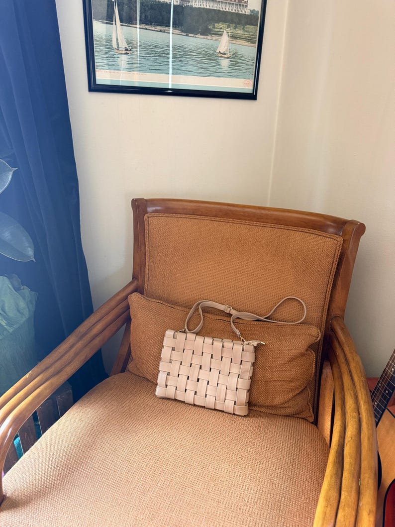 Pu&ograve; includere: Una borsa a mano beige chiaro intrecciata con una tracolla &egrave; appoggiata su un cuscino marrone. La borsa ha una forma rettangolare e una chiusura a cerniera. La borsa &egrave; su una sedia marrone con schienale e braccioli intrecciati.