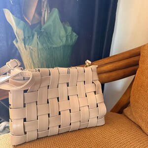 Pu&ograve; includere: Una borsa beige intrecciata con tracolla &egrave; appoggiata su una sedia. La borsa presenta un design a trama a cestino, con strisce rettangolari di materiale intrecciate tra loro. La borsa &egrave; di colore neutro e la tracolla &egrave; fissata con hardware color oro.