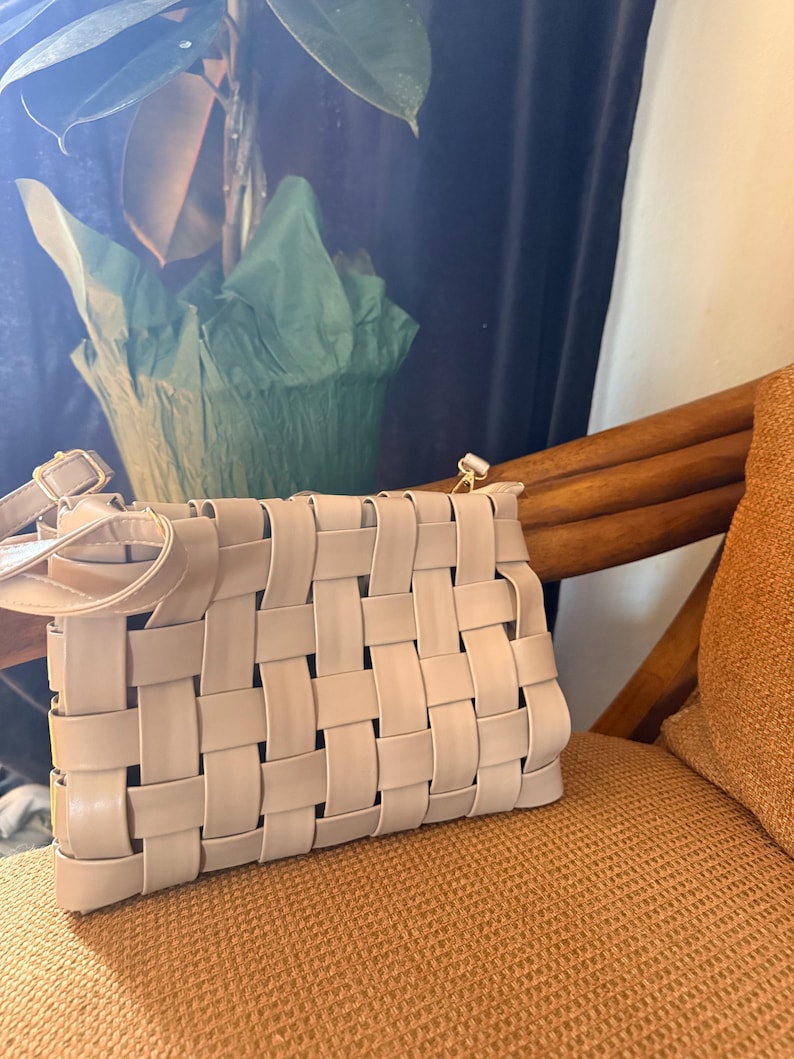 Pu&ograve; includere: Una borsa beige intrecciata con tracolla &egrave; appoggiata su una sedia. La borsa presenta un design a trama a cestino, con strisce rettangolari di materiale intrecciate tra loro. La borsa &egrave; di colore neutro e la tracolla &egrave; fissata con hardware color oro.