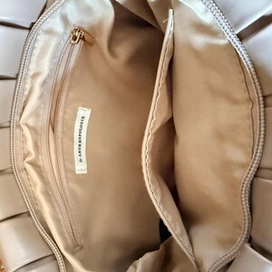 Pu&ograve; includere: Borsa beige con design intrecciato all'esterno. L'interno &egrave; beige, con una tasca con cerniera e un'etichetta "by ANTHROPOLOGIE". La borsa ha una cerniera dorata e una forma rettangolare.