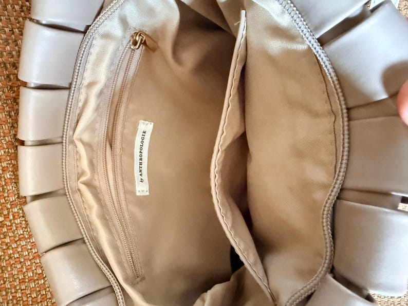 Pu&ograve; includere: Borsa beige con design intrecciato all'esterno. L'interno &egrave; beige, con una tasca con cerniera e un'etichetta "by ANTHROPOLOGIE". La borsa ha una cerniera dorata e una forma rettangolare.