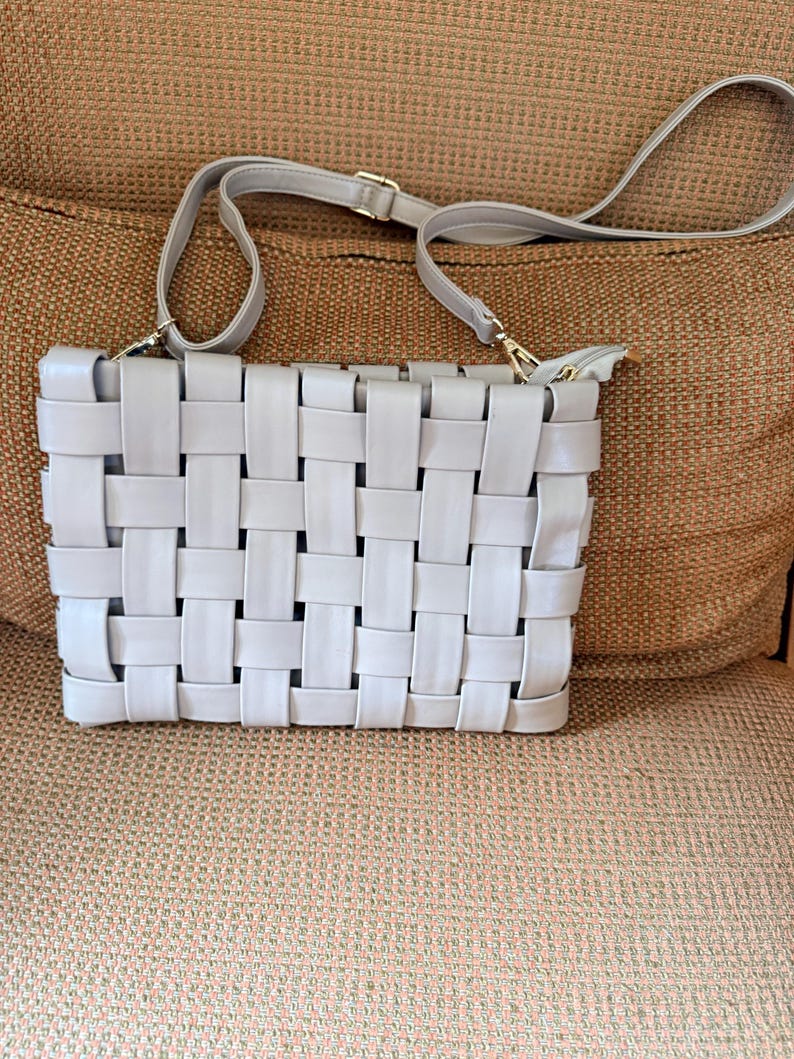 Pu&ograve; includere: Una borsa a tracolla grigio chiaro con un design intrecciato. La borsa ha una forma rettangolare, una lunga tracolla regolabile e hardware color oro. Il motivo intrecciato crea una superficie strutturata.
