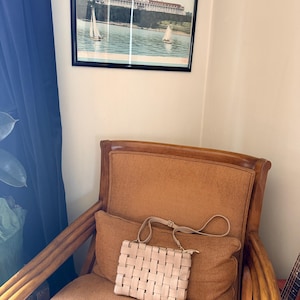 Pu&ograve; includere: Una borsa a mano rosa chiaro intrecciata con una tracolla &egrave; appoggiata su un cuscino marrone. La borsa ha una forma rettangolare con un design intrecciato. Una foto incorniciata di un edificio su un lago &egrave; appesa al muro.