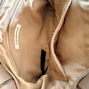 Pu&ograve; includere: Vista interna di una borsa beige con pi&ugrave; scomparti. La borsa presenta una tasca con cerniera e una tasca a bustina. Una piccola etichetta all'interno della borsa recita "& ANTHROPOLOGIE". L'interno della borsa &egrave; di colore beige chiaro.