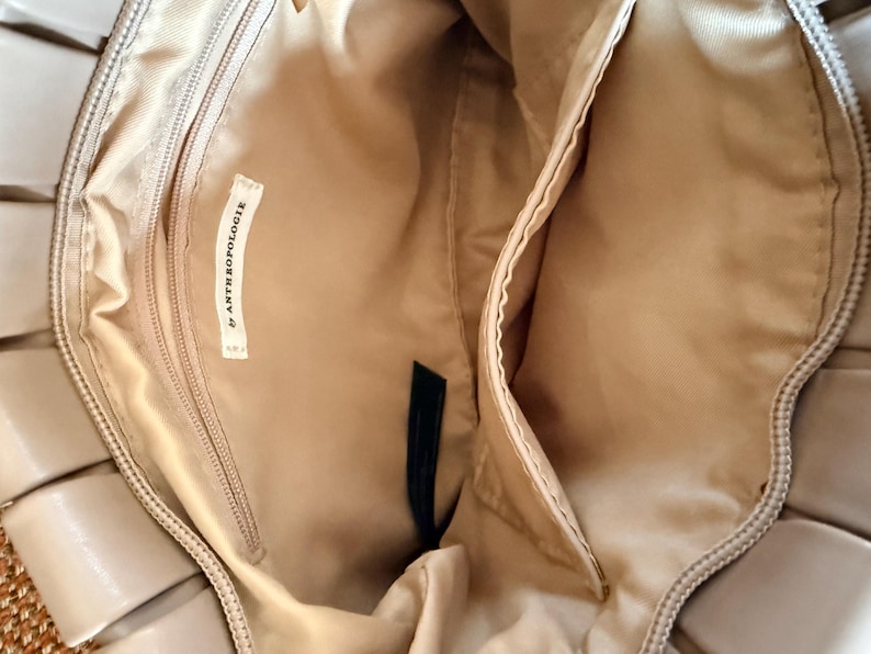 Pu&ograve; includere: Vista interna di una borsa beige con pi&ugrave; scomparti. La borsa presenta una tasca con cerniera e una tasca a bustina. Una piccola etichetta all'interno della borsa recita "& ANTHROPOLOGIE". L'interno della borsa &egrave; di colore beige chiaro.