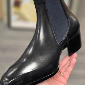 May include: A black leather Chelsea boot with a smooth, polished finish, a pointed toe, a stacked block heel, and a dark navy blue elastic side panel. The boot features a sleek silhouette with clean lines and a structured, high-ankle design.
