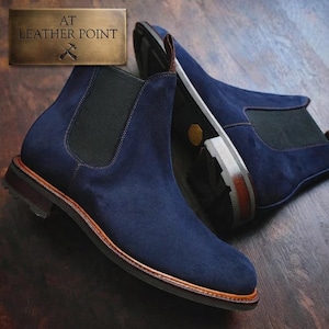May include: A pair of navy blue suede Chelsea boots with black elastic side panels and a brown leather welt. The boots feature a stacked heel and a textured outsole. A metallic plaque in the corner displays the text AT LEATHER POINT.