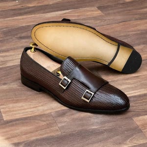 May include: Brown leather dress shoes with a woven texture and double buckle detail. One shoe is upright, the other is upside down, revealing the sole. The shoes are on a wooden surface.