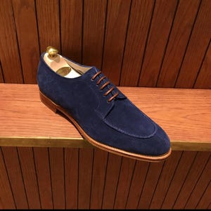May include: A navy blue suede dress shoe with brown leather trim and brown laces. The shoe is displayed on a wooden shelf with a shoe tree inside. The shoe has a classic design.